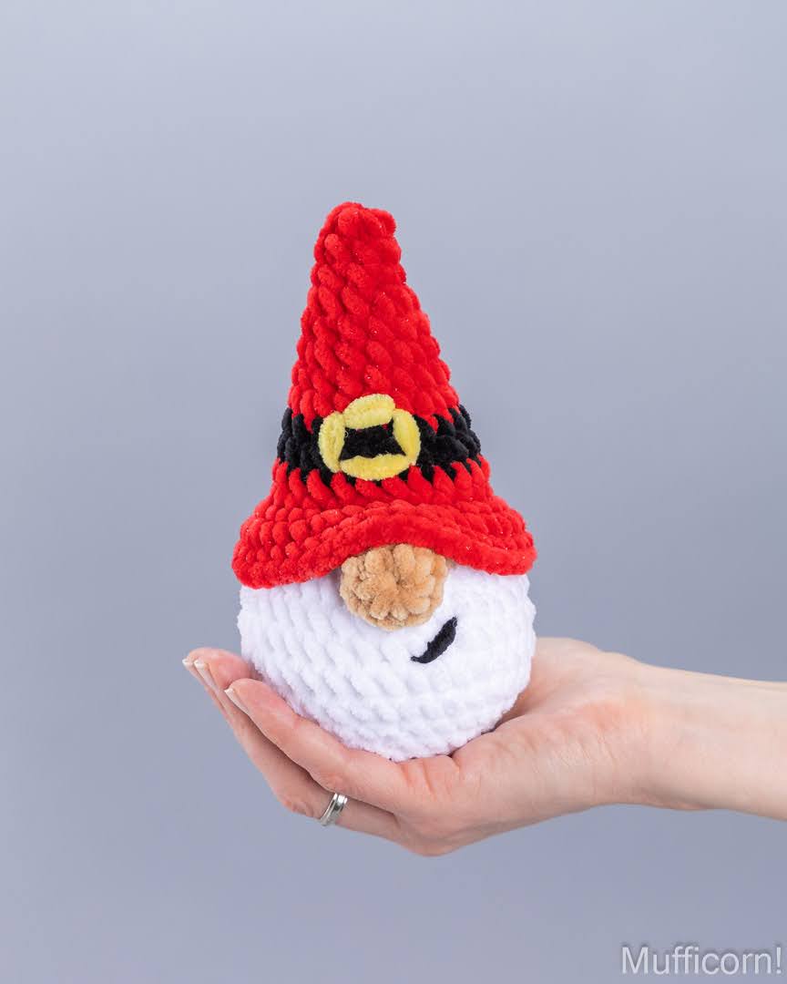 A hand holds a small crocheted gnome doll with a red pointed hat, black band, yellow circular detail, white body, and brown nose against a plain gray background.