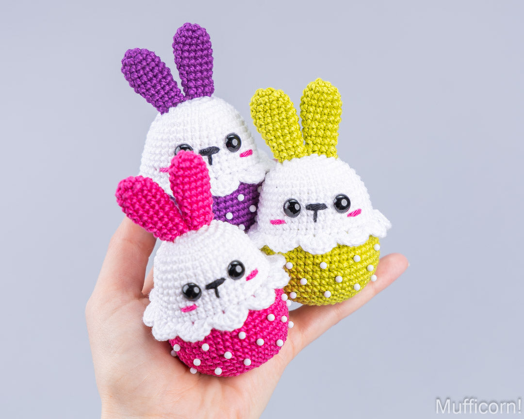 Three handmade crocheted bunny amigurumi dolls are held in a person's hand against a plain light gray background. The bunnies have white faces and bodies, with purple, pink, and yellow ears, and are decorated with colorful polka-dot patterns.
