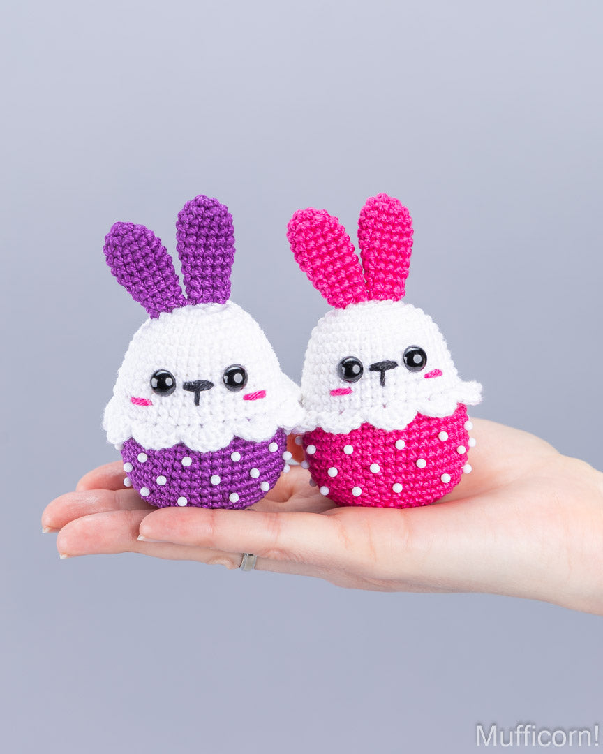 Two small crocheted bunny cupcake toys are held in a person's hand against a plain light gray background. One bunny is white with purple ears and a purple polka-dot skirt, and the other is white with pink ears and a pink polka-dot skirt.