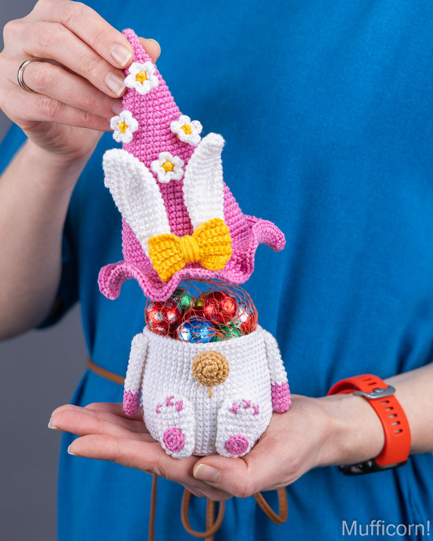 A person in a blue shirt holds a crocheted Easter bunny ornament shaped like a candy jar, with a pink hat, white bunny body, yellow bow, and colorful chocolate candies inside. The photo is cropped to the person's hands and torso against a neutral gray background.