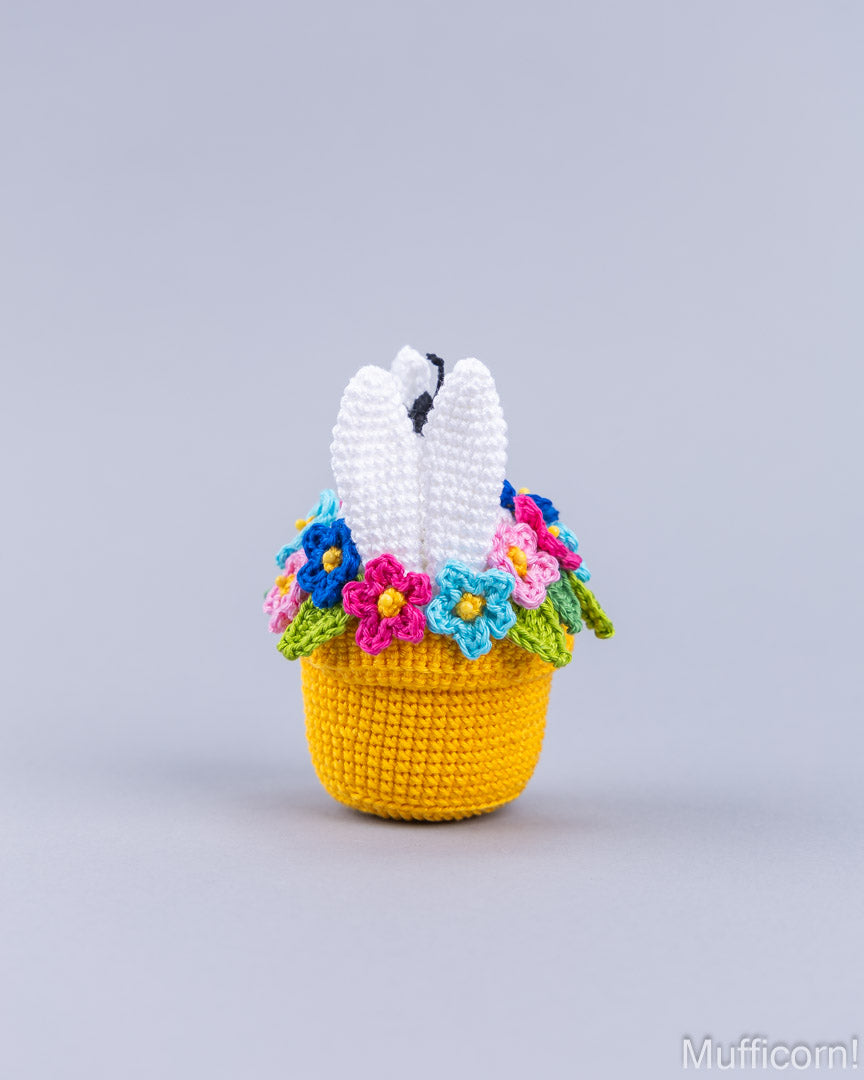 A crocheted Easter basket with a white bunny inside and colorful flowers around the rim, set against a solid light gray background.