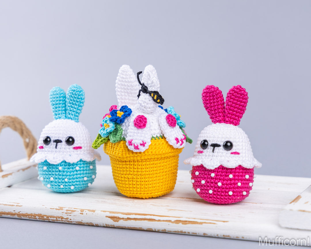 Three crocheted bunny-shaped ornaments displayed on a white rustic wooden surface against a light gray background. The left bunny is light blue and white, the center bunny is yellow and white with colorful flowers, and the right bunny is pink and white with white polka dots.