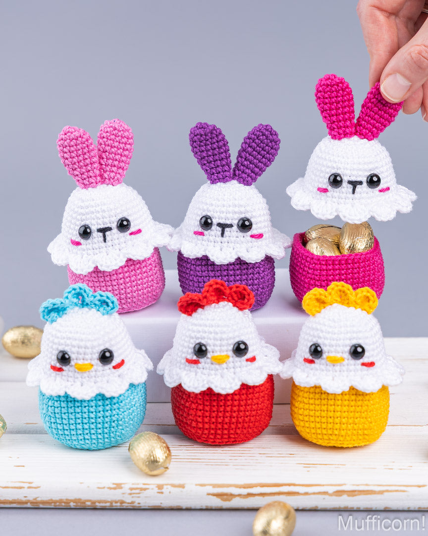A set of crocheted Easter-themed amigurumi figures, including bunnies and chicks, in pink, purple, blue, red, and yellow, arranged on a white wooden surface with gold chocolate eggs. A hand is visible holding one bunny's ear against a gray background.