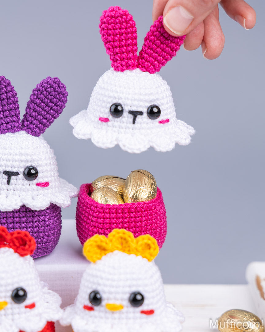 Handcrafted crocheted Easter-themed bunny and chick figures with colorful ears and beaks, alongside a pink basket filled with gold foil chocolate eggs, shown against a soft gray background.