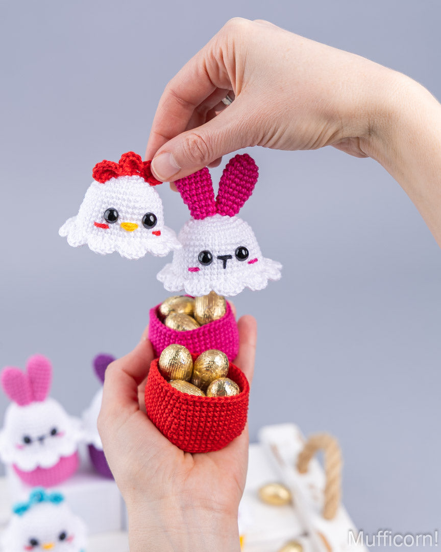 A person's hands holding a small crocheted red basket filled with gold foil chocolate eggs, with two small crocheted bunny and chick character toppers above the basket against a soft gray studio background.