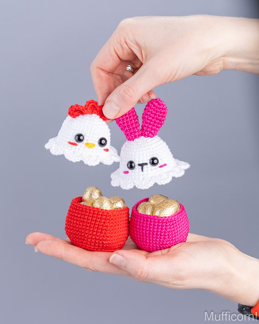 A pair of hands holding crocheted Easter-themed amigurumi toys against a solid gray background. The set includes a white chicken with a red comb, a white bunny with pink ears, and two small red and pink pots filled with gold-colored chocolate eggs.