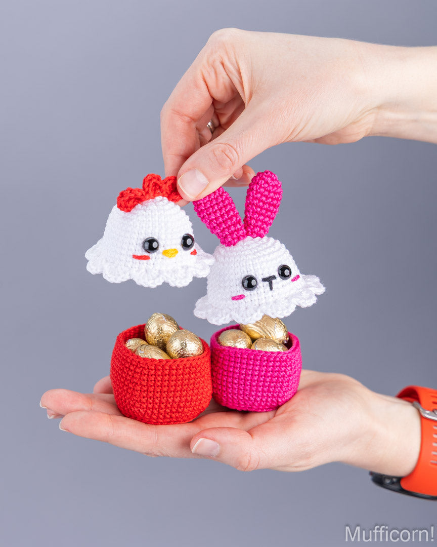 A person's hands holding two small crocheted Easter-themed baskets filled with gold foil eggs. One basket is red with a white chick decoration on top, and the other is pink with a white bunny decoration on top, set against a solid gray background.