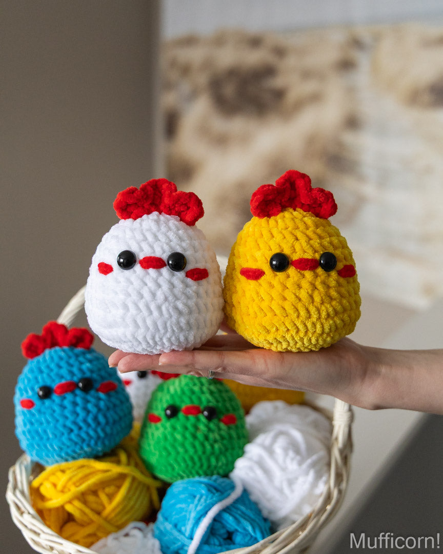A hand holds two knitted amigurumi chicks, one white and one yellow, both with red crowns and black eyes. A wicker basket filled with colorful yarn balls sits in the background.
