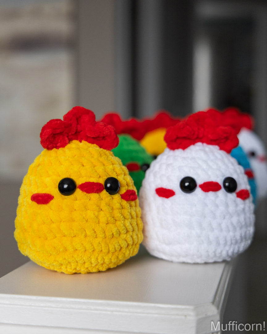 Two crocheted chicken toys in yellow and white with red combs and red beaks, sitting on a white surface with a blurred indoor background. A Mufficorn logo appears in the bottom right corner.