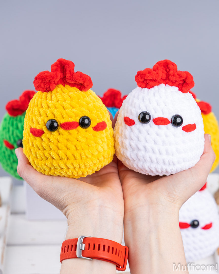 Two small crocheted chicken plush toys held in a person's hands against a soft gray background. One toy is yellow with a red comb and black eyes, and the other is white with a red comb and red nose.
