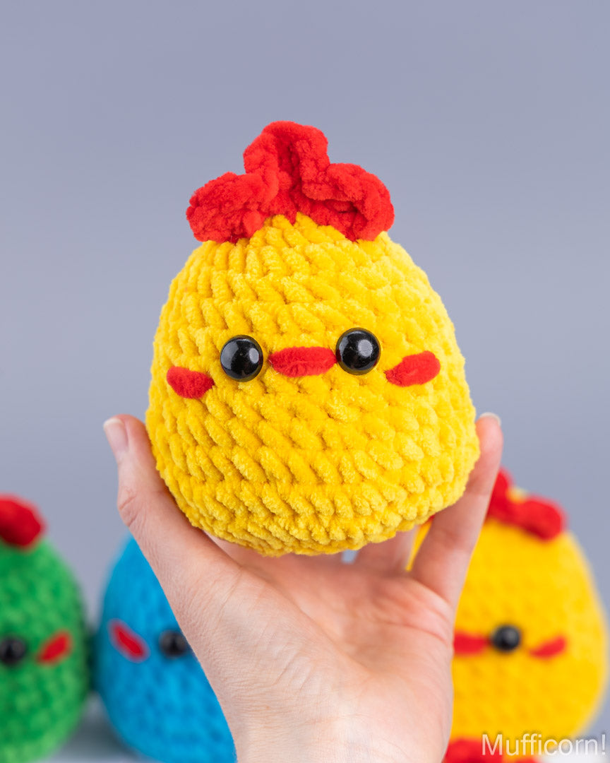 A hand holds a yellow crocheted chick toy with a red crest, black bead eyes, and red stitched cheeks against a soft gray background, with other colorful chick toys blurred in the background.