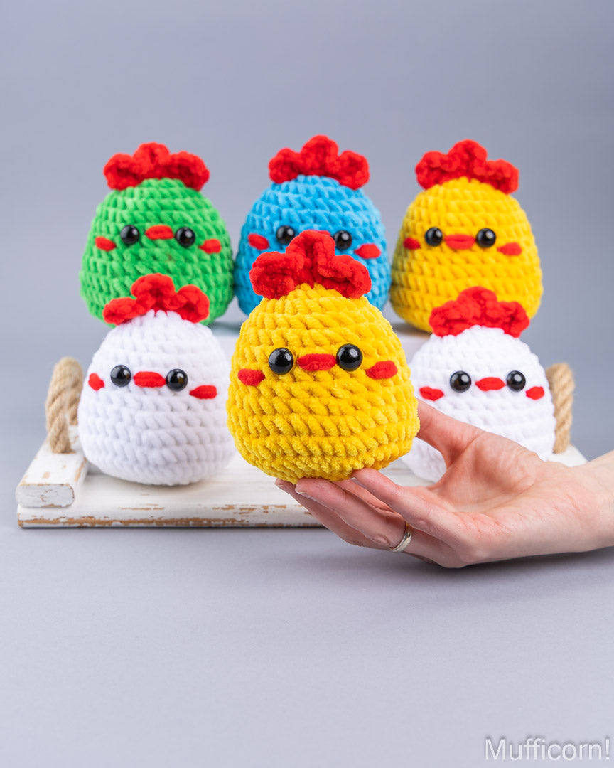 A hand holds a yellow crocheted chick toy in the foreground, with five more colorful chick toys arranged behind it on a small white tray against a light gray background.