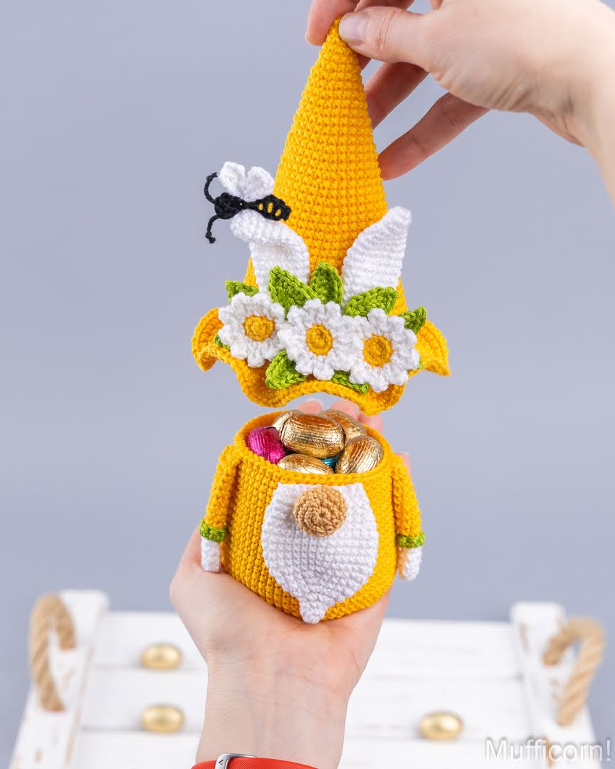 A crocheted Easter gnome ornament held in a hand, with its hat being lifted by another hand. The gnome is yellow and white with white daisies, green leaves, a small black and yellow bee, and gold chocolate eggs inside its pouch.