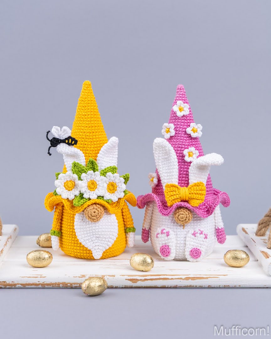 Two crocheted Easter garden gnome dolls displayed on a white wooden surface with gold eggs. One gnome is yellow with a white beard, white bunny ears, daisy flowers, and a black-and-yellow bumblebee accessory.