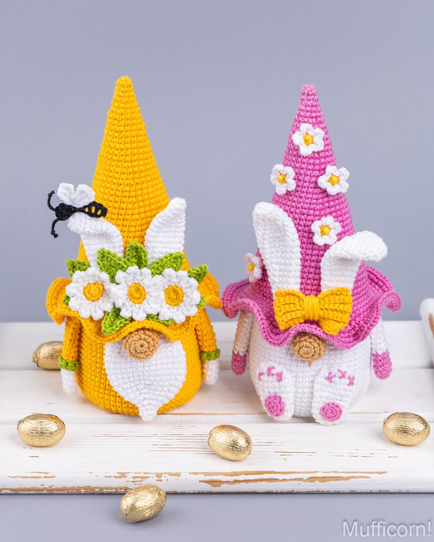 Two crocheted Easter gnomes displayed on a white wooden surface with scattered gold chocolate eggs. The left gnome is yellow and white with a flower crown and black spider detail.