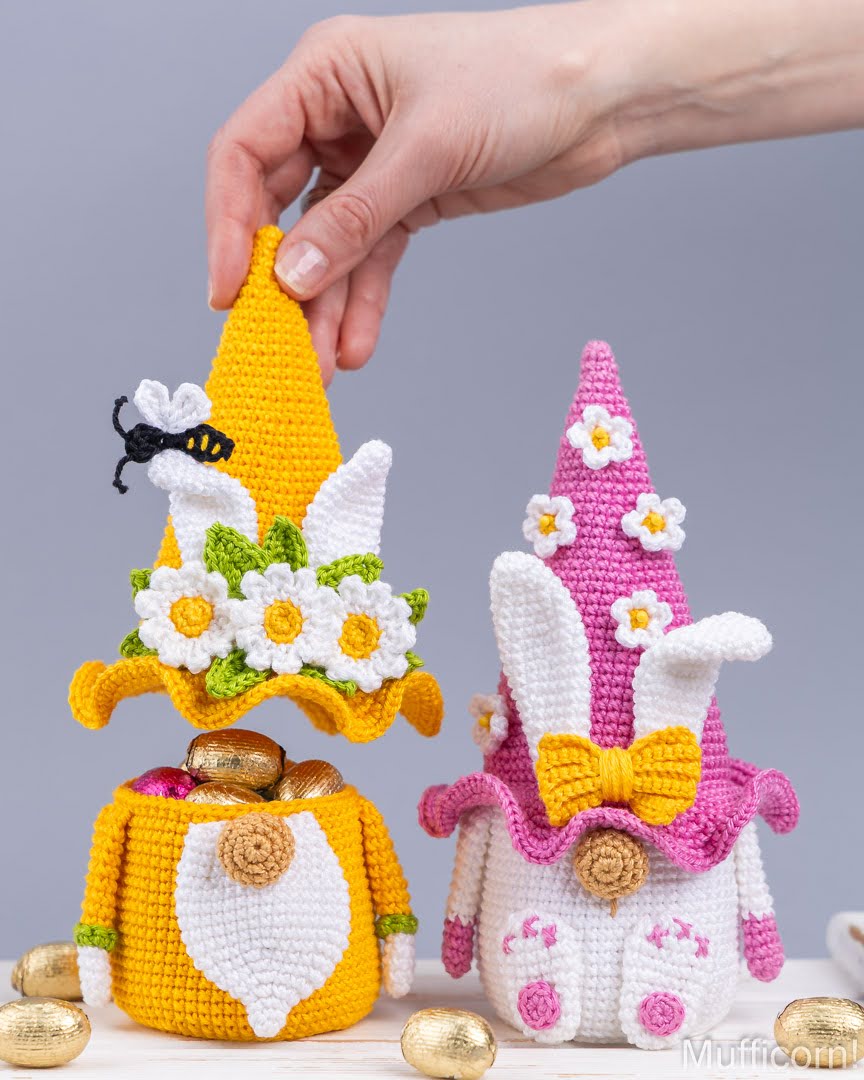 Two crocheted Easter-themed gnomes with pointed hats, one yellow and one pink, displayed on a white surface with gold chocolate eggs. A human hand is adjusting the yellow gnome's hat, set against a solid gray studio background.