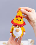 A crocheted gnome chicken candy keeper  being held by two hands, filled with gold foil chocolates. The item has a yellow body, white belly, red ruffled collar and hat, and small black facial details.