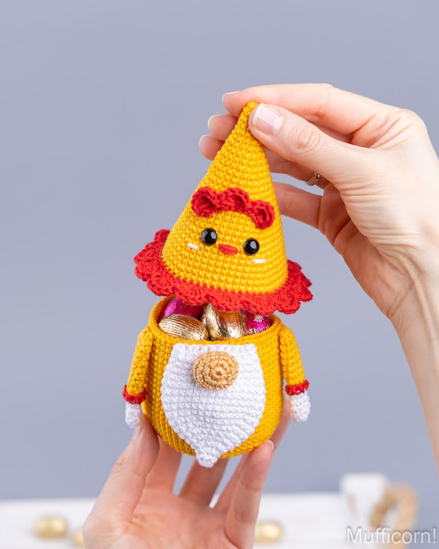 A crocheted gnome chicken candy keeper  being held by two hands, filled with gold foil chocolates. The item has a yellow body, white belly, red ruffled collar and hat, and small black facial details.