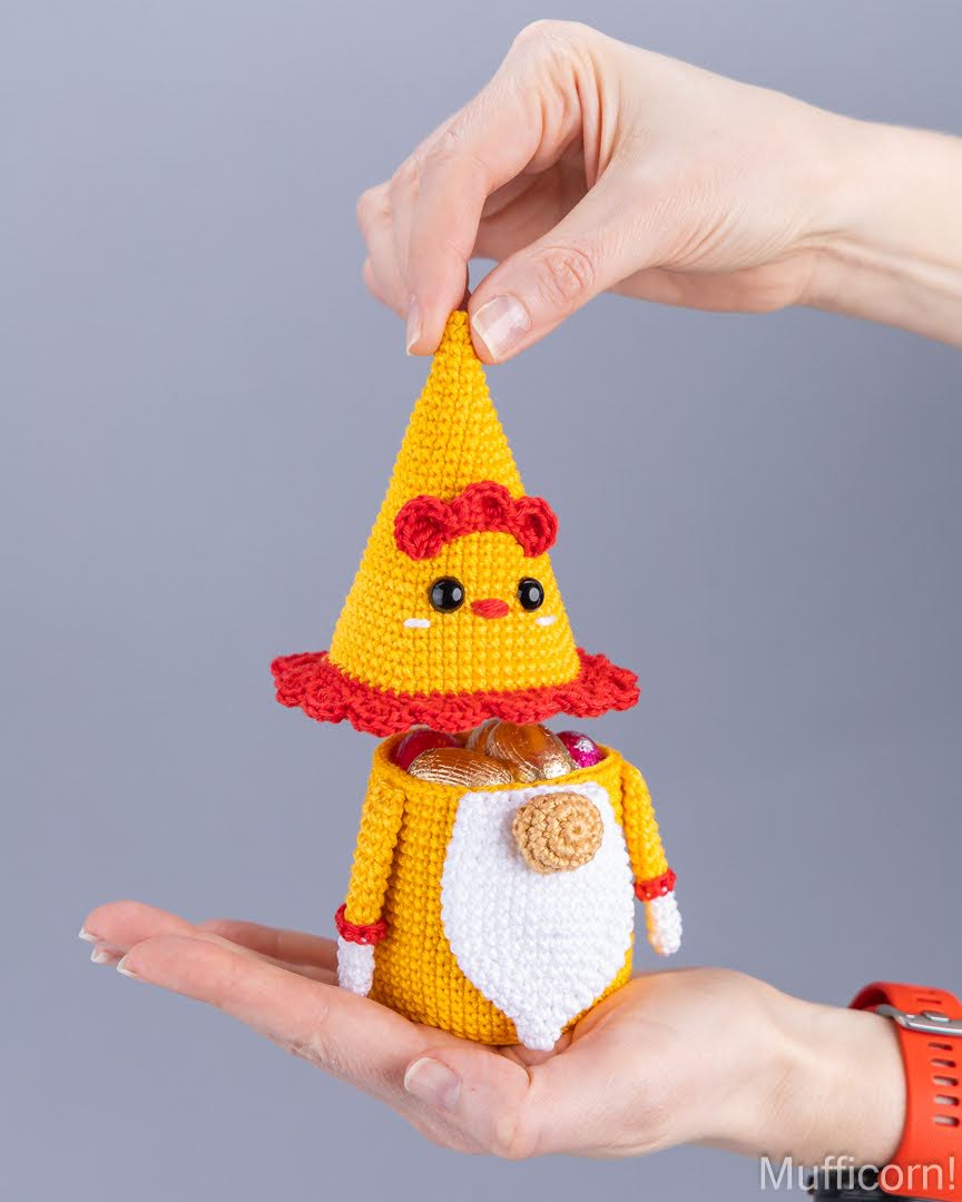 A crocheted gnome chicken candy keeper with a yellow body, white beard, red hat trim, and small brown hat on its head, being held in two hands against a plain gray background.