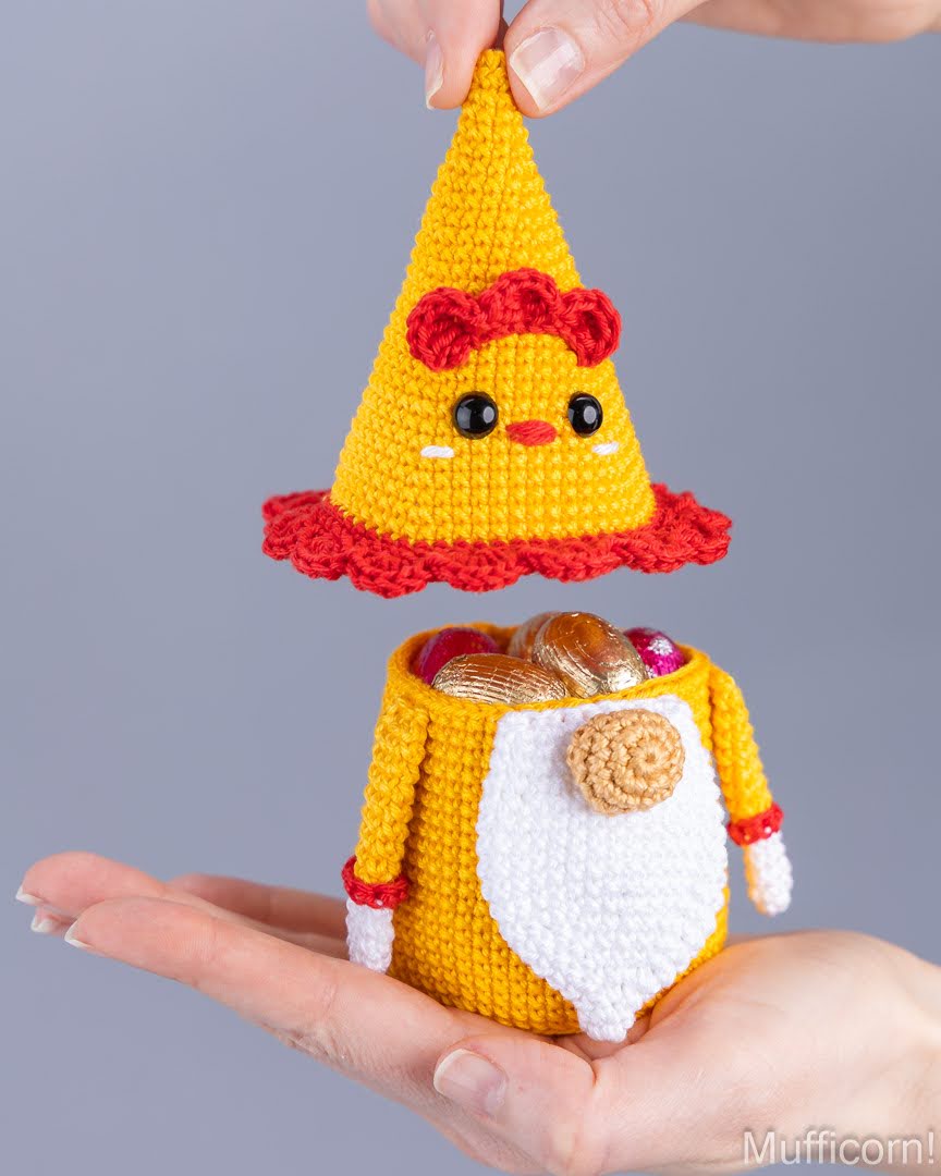 A crocheted amigurumi character ornament or pouch is held between two hands against a plain gray studio background. The top hat-like piece is yellow with red trim and a face, while the lower pouch-like body is also yellow with a white beard and small red accents.