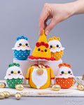 A hand is placing a crocheted Easter-themed ornament on top of a small display of colorful handmade chick and egg decorations, with gold chocolate eggs scattered around on a light gray background.