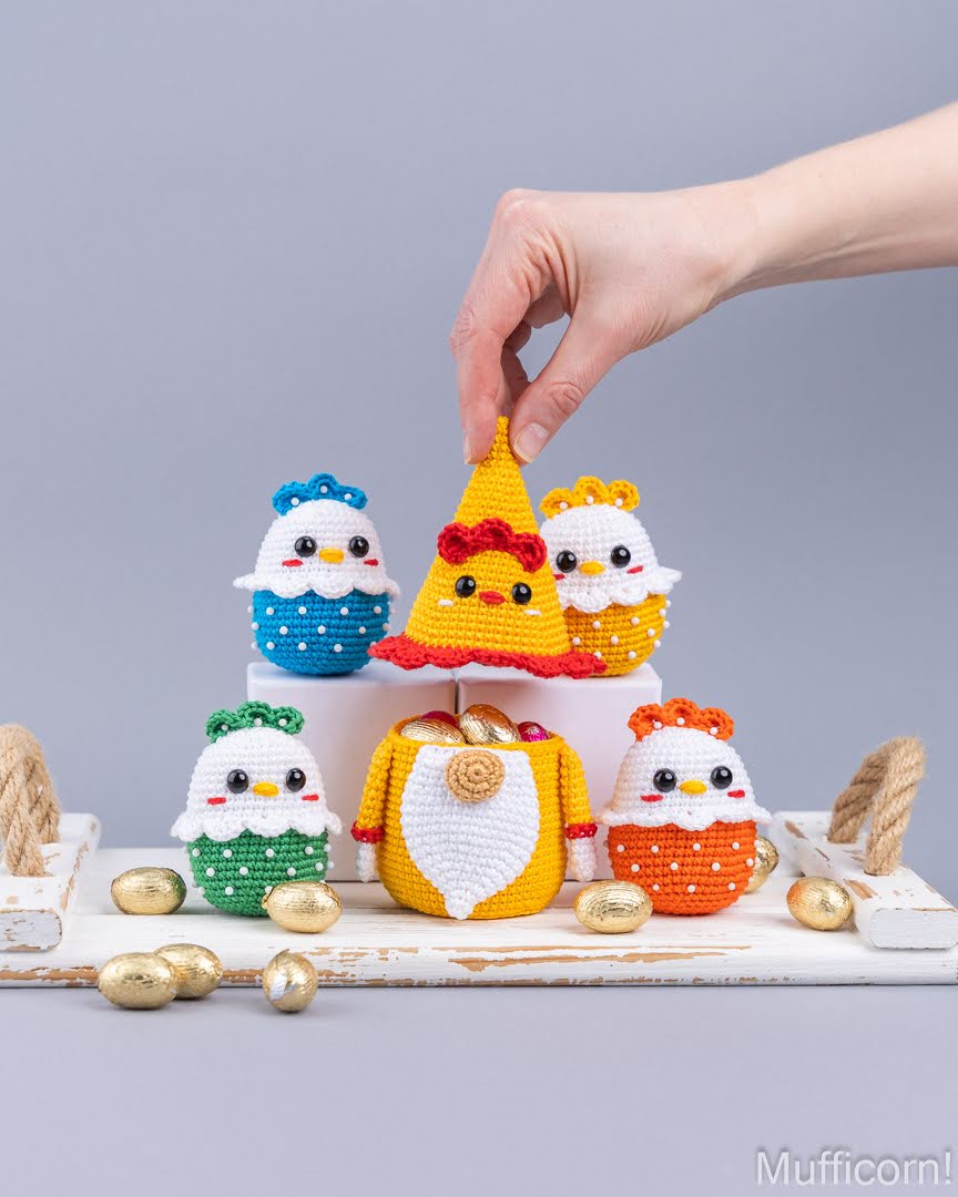 Five crocheted Easter egg characters in blue, green, yellow, orange, and red are displayed on a white wooden stand, with a hand placing the central yellow character. Small gold chocolate eggs are scattered around on a light gray background.