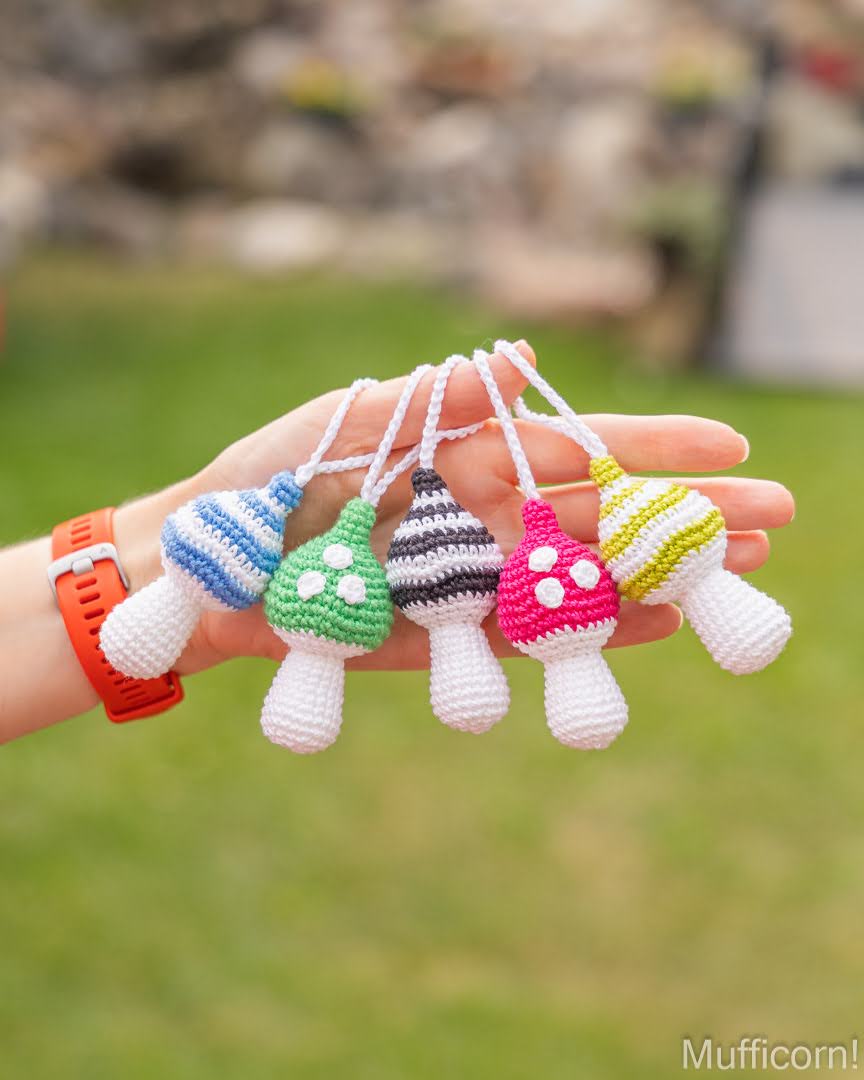 A hand holds five small crocheted mushroom-shaped ornaments in different colors, including blue, green, black and white, pink, and yellow and white, against a blurred green outdoor background.
