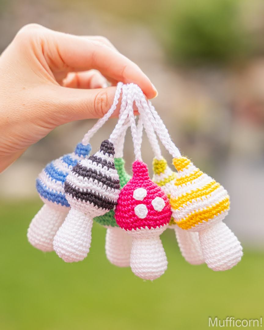 A hand holds a cluster of small crocheted mushroom-shaped accessories in different color patterns, including blue and white, black and white, green and white, pink and white, and yellow and white, against a blurred green outdoor background.