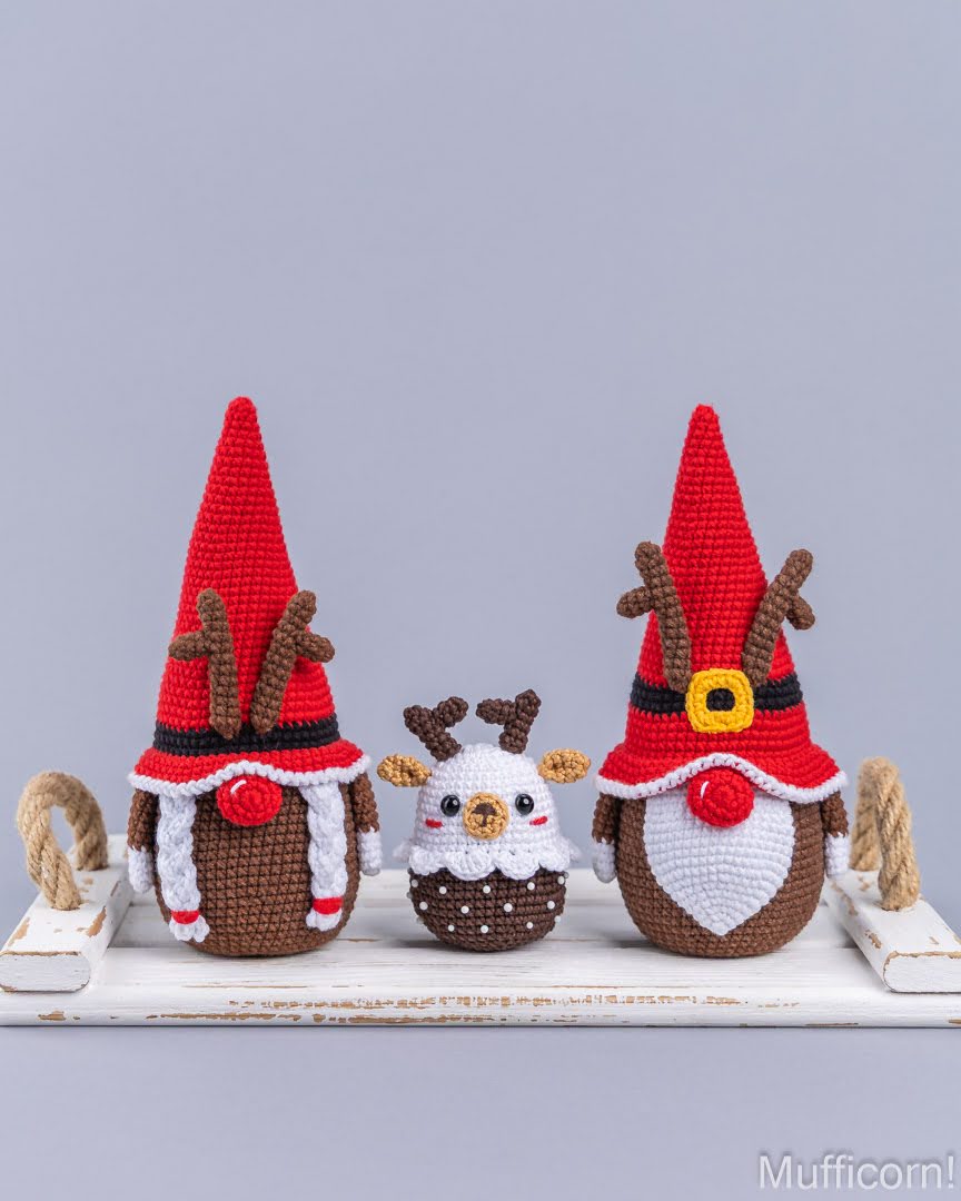 Three crocheted Christmas-themed amigurumi figures displayed on a white wooden shelf against a light gray background. The set includes two reindeer gnomes with red pointed hats, brown antlers, white beards, and red noses, and one small white and brown reindeer figure in the center.
