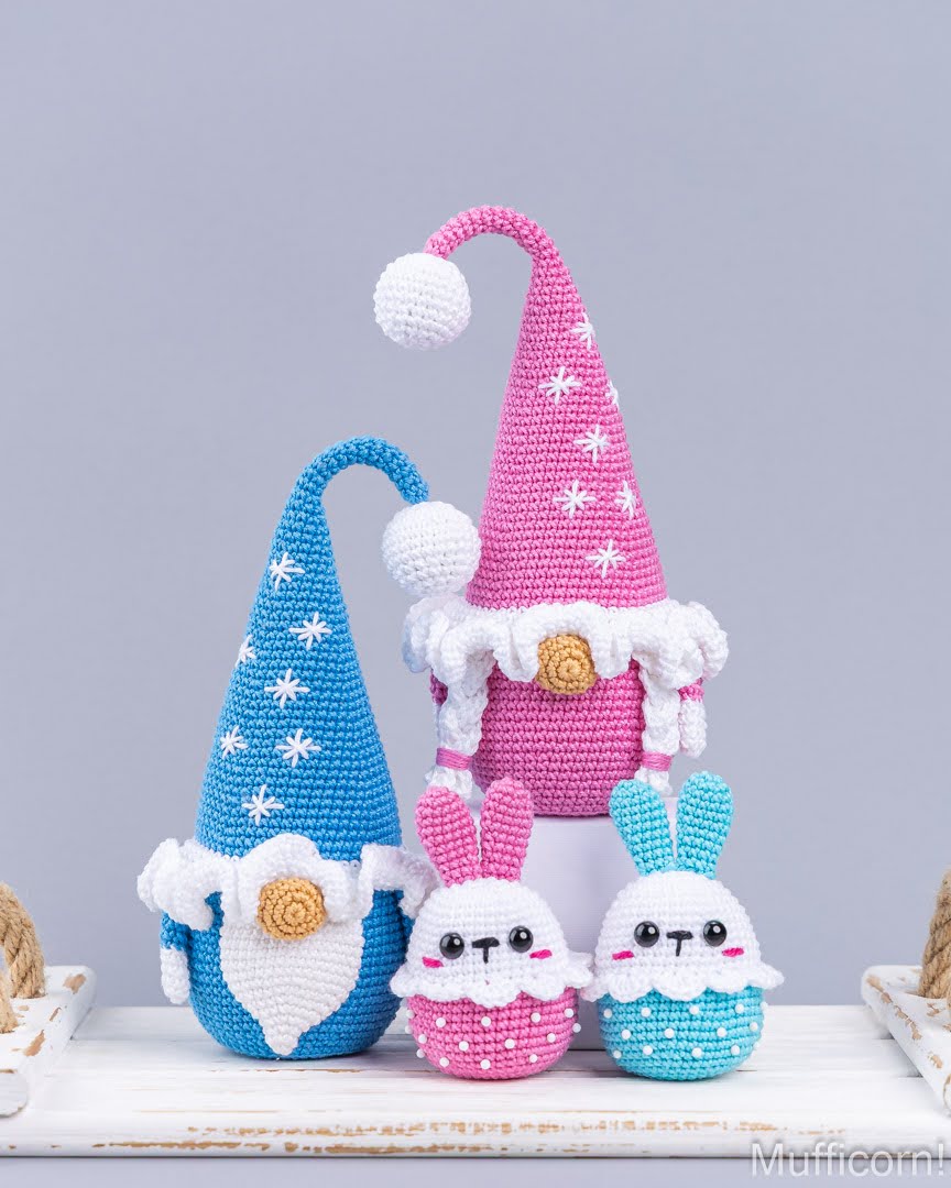 Three crocheted Easter-themed amigurumi figures: a blue gnome with a white beard and hat, a pink gnome in a pink hat, and two small bunny figures in pink and blue, displayed on a white wooden surface against a gray background.