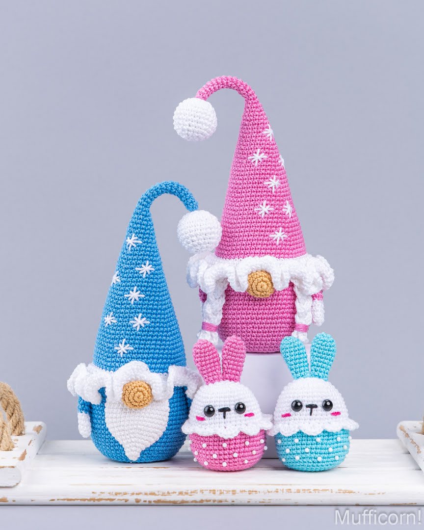 Three crocheted Easter-themed decorative figures on a white shelf against a light gray background: a tall blue gnome with white star pattern and beard, a tall pink gnome with white trim and pom-pom, and two small bunny cupcake figures in pink and blue.