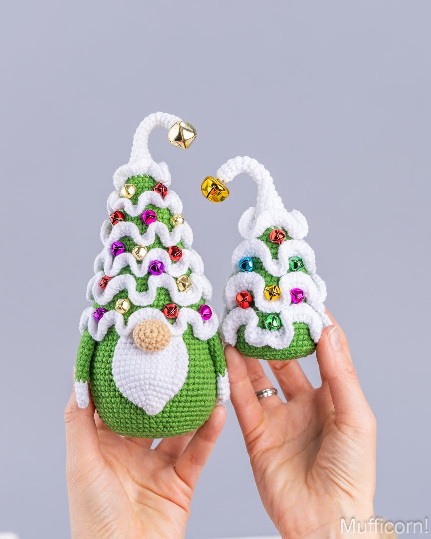 Two crocheted Christmas gnome ornaments shaped like small trees, held in both hands against a plain light gray background. The larger ornament is green with white wavy trim, multicolored rhinestones, a gold bell, and a white beard with a beige nose.