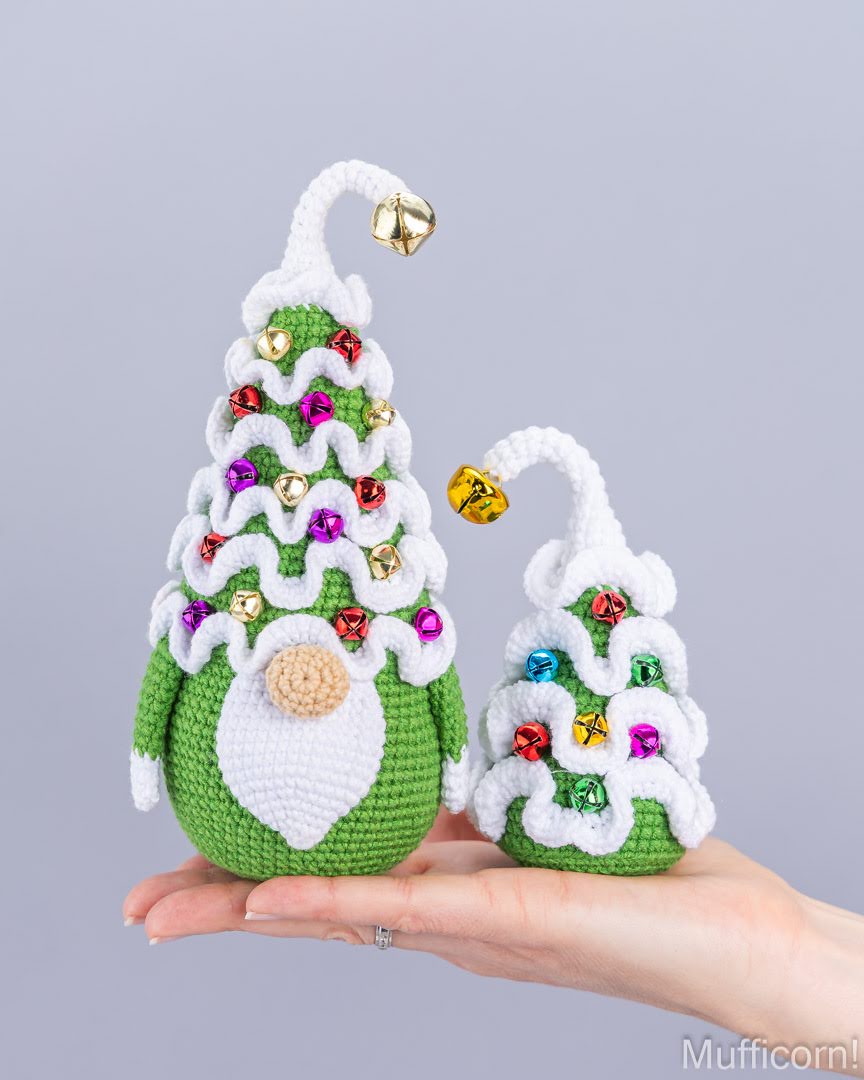 Two crocheted Christmas tree ornaments with gnome heads are displayed on a person's hand against a solid gray background. The larger ornament is green with white trim, colorful rhinestone decorations, and a gold bell.