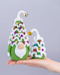 A hand holds two crocheted Christmas tree gnomes in green and white, decorated with colorful rhinestones and gold bells, against a plain light gray background.
