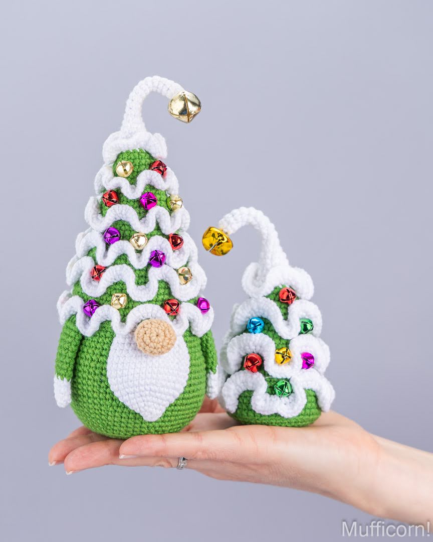 A hand holds two crocheted Christmas tree gnomes in green and white, decorated with colorful rhinestones and gold bells, against a plain light gray background.