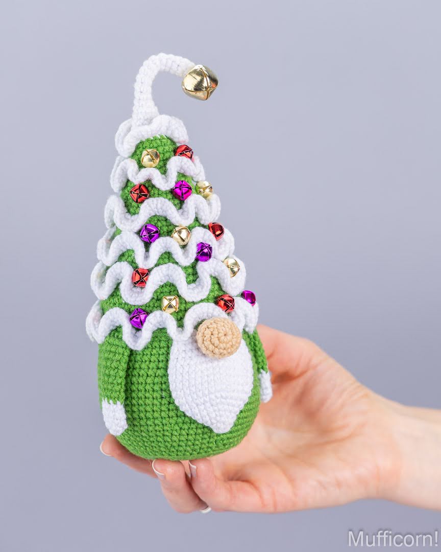 A hand holding a crocheted Christmas gnome ornament shaped like a tree, decorated with white ruffles, colorful rhinestones, a green body, and a small gold bell on a light gray background.