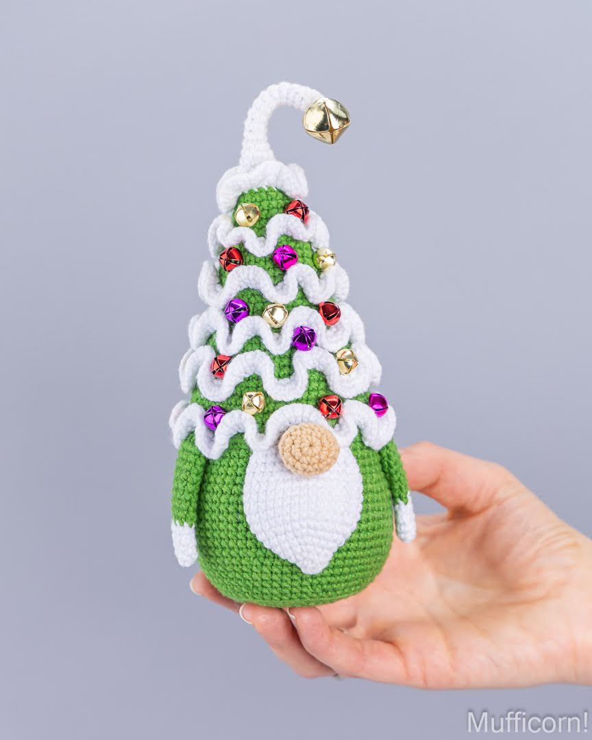A hand holds a crocheted Christmas gnome ornament shaped like a decorated Christmas tree, with green and white yarn, small shiny ornaments, and a gold bell on a plain light gray background.