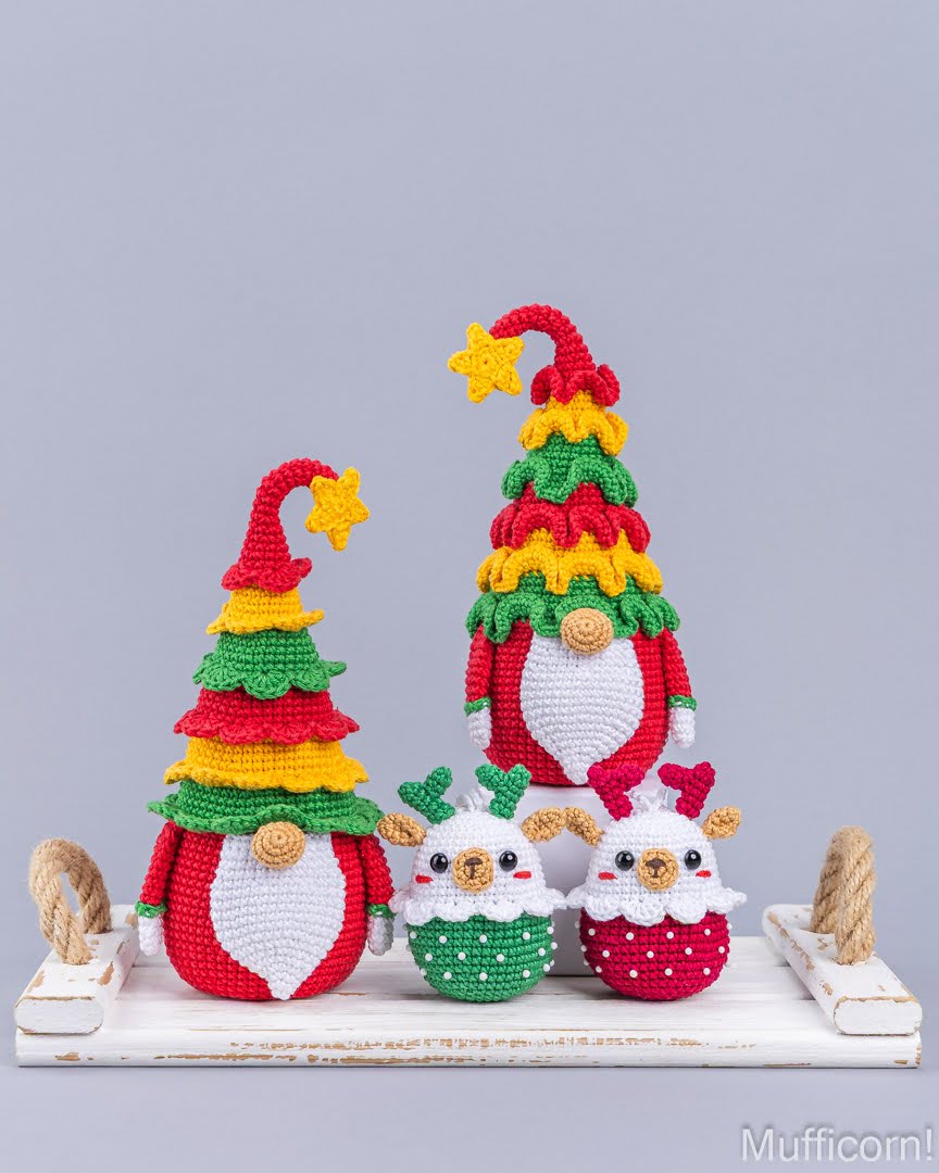 Three crocheted Christmas gnome figures displayed on a white wooden shelf against a light gray background. The set includes two tall gnomes styled like Christmas trees with red, yellow, and green layers and two smaller gnomes dressed as Santa and elf in red and green outfits.