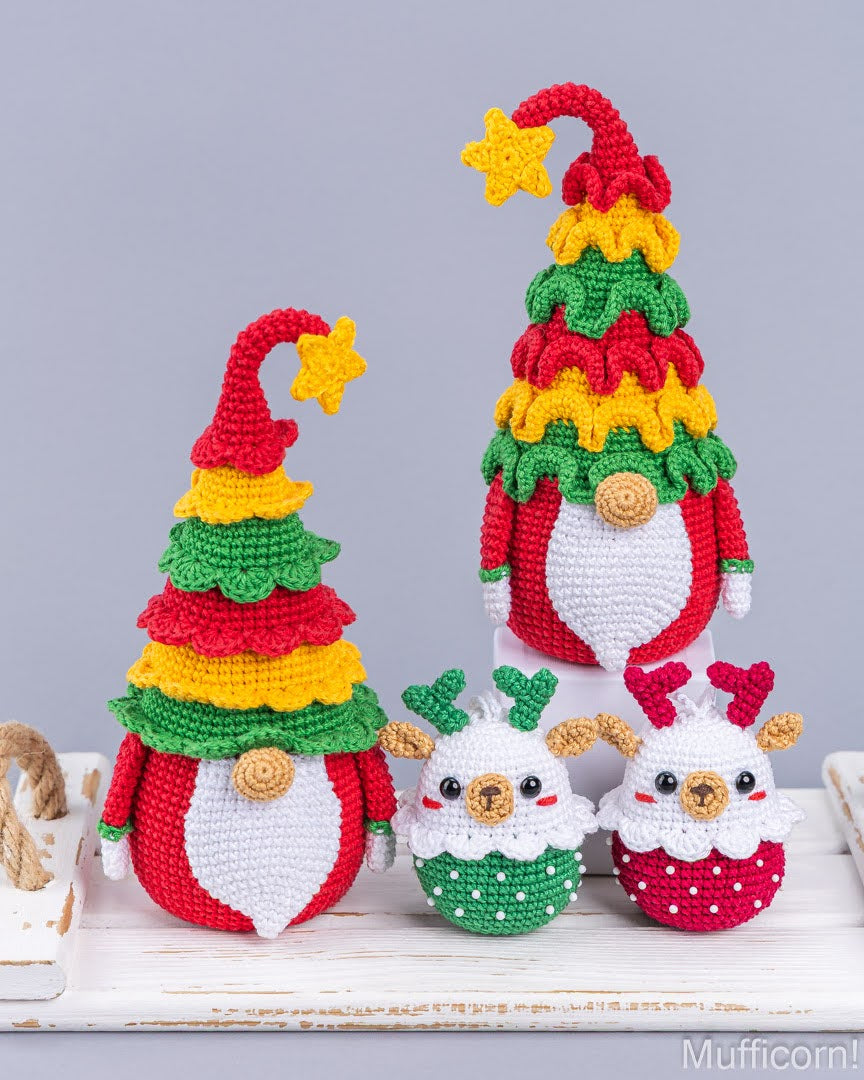 Three crocheted Christmas-themed gnomes and two small reindeer ornaments arranged on a white wooden surface against a light gray background. The gnomes have red and white bodies, multicolored tree-like hats, and yellow star accents, while the reindeer are white with red and green details.