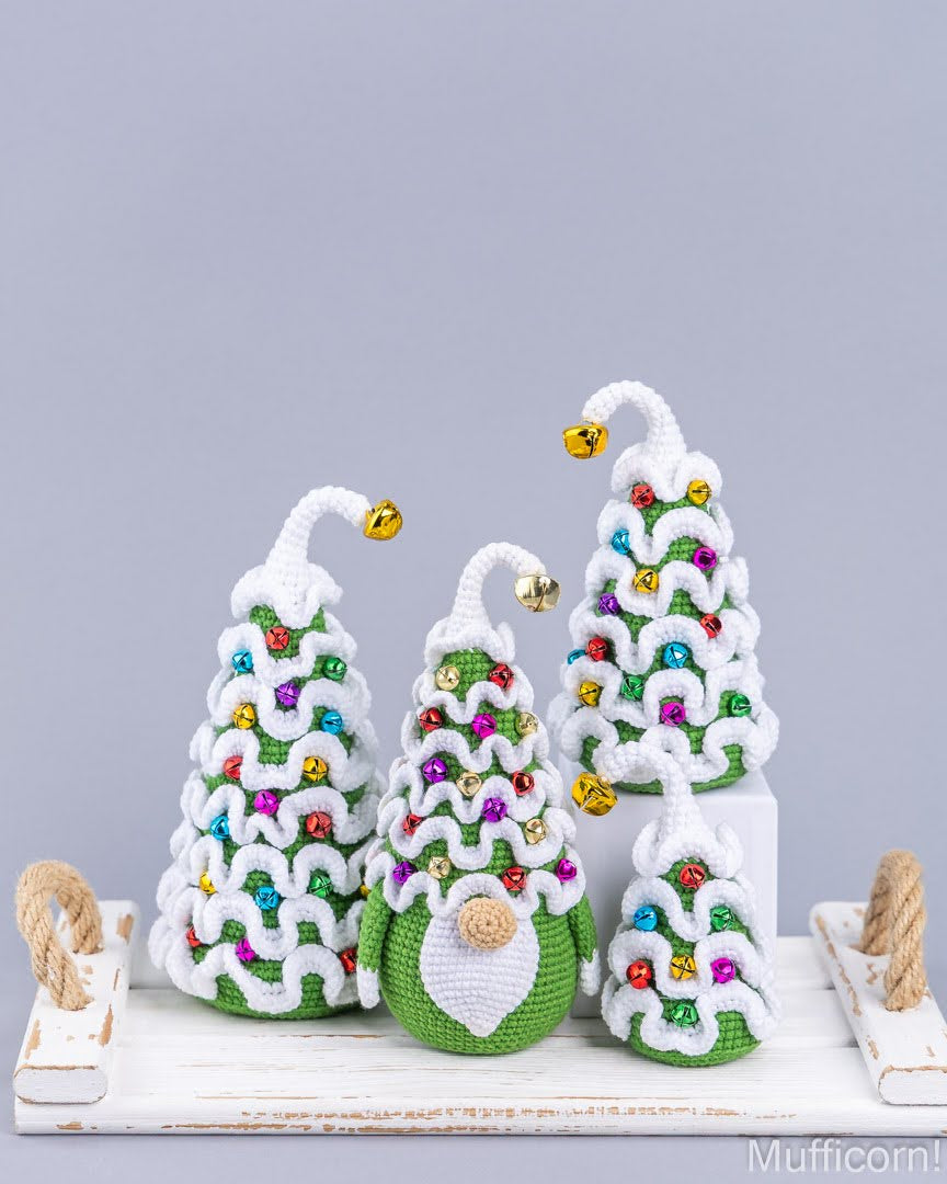 Four crocheted Christmas tree ornaments in different sizes, decorated with colorful beads and gold bells, arranged on a white wooden tray against a solid gray background.