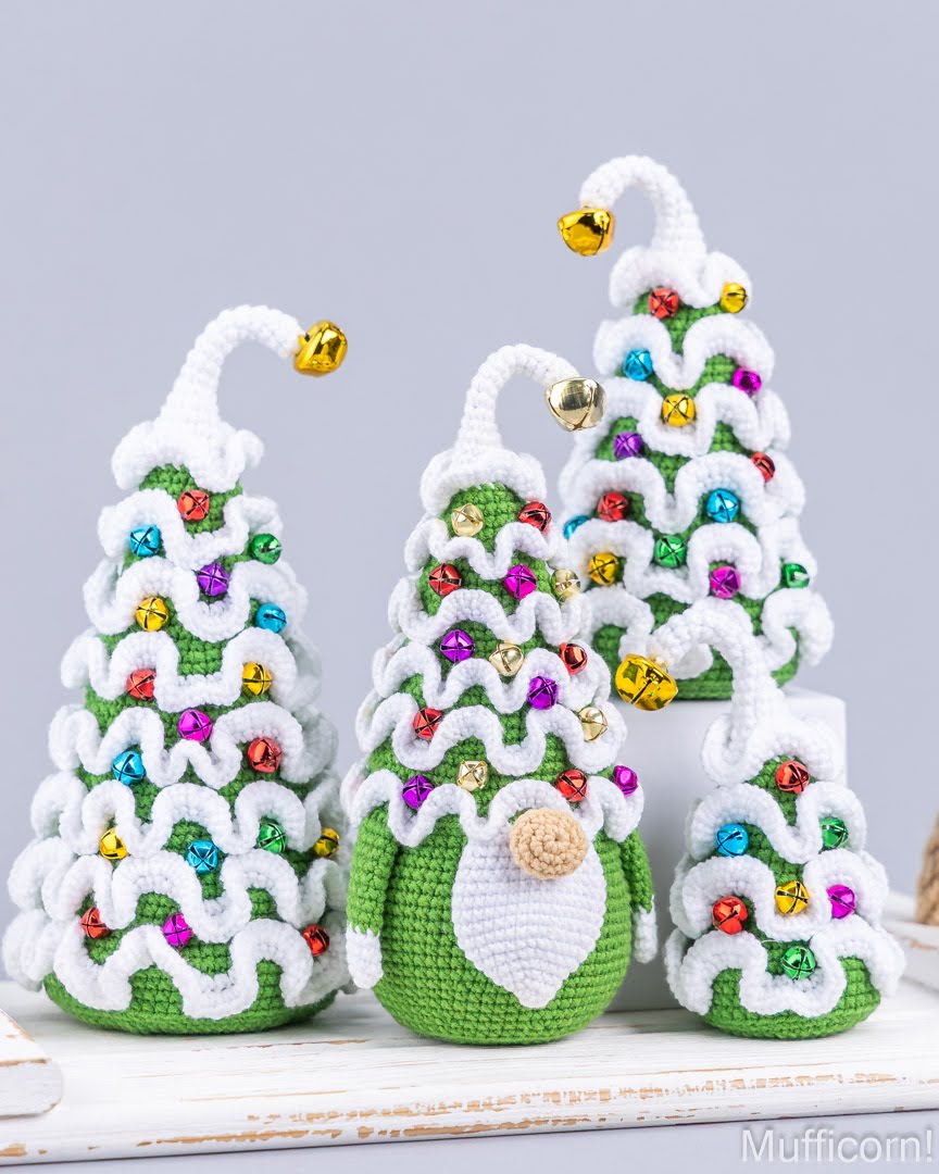 Three crocheted Christmas tree gnomes in green and white, decorated with colorful rhinestones and gold bells, arranged on a white surface against a light gray background.