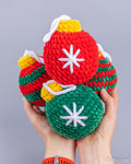 Three hand-crocheted Christmas ornament toys in red, green, and striped red/green colors, held in two hands against a solid gray background.