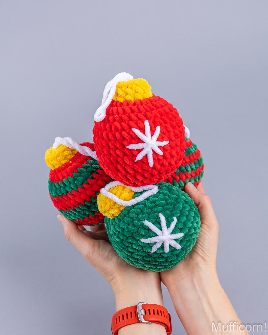 Three crocheted Christmas ornament-shaped items in red, green, and yellow are held in two hands against a plain gray background. Each ornament has a white star detail and a white hanging loop.