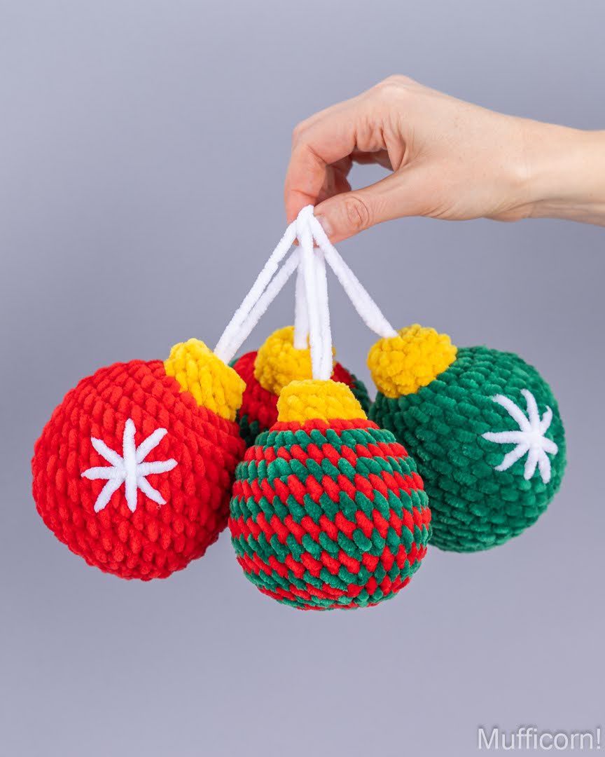 Three knitted Christmas ornament-shaped toys in red, green, and red/green striped patterns, held by a hand against a plain gray background.