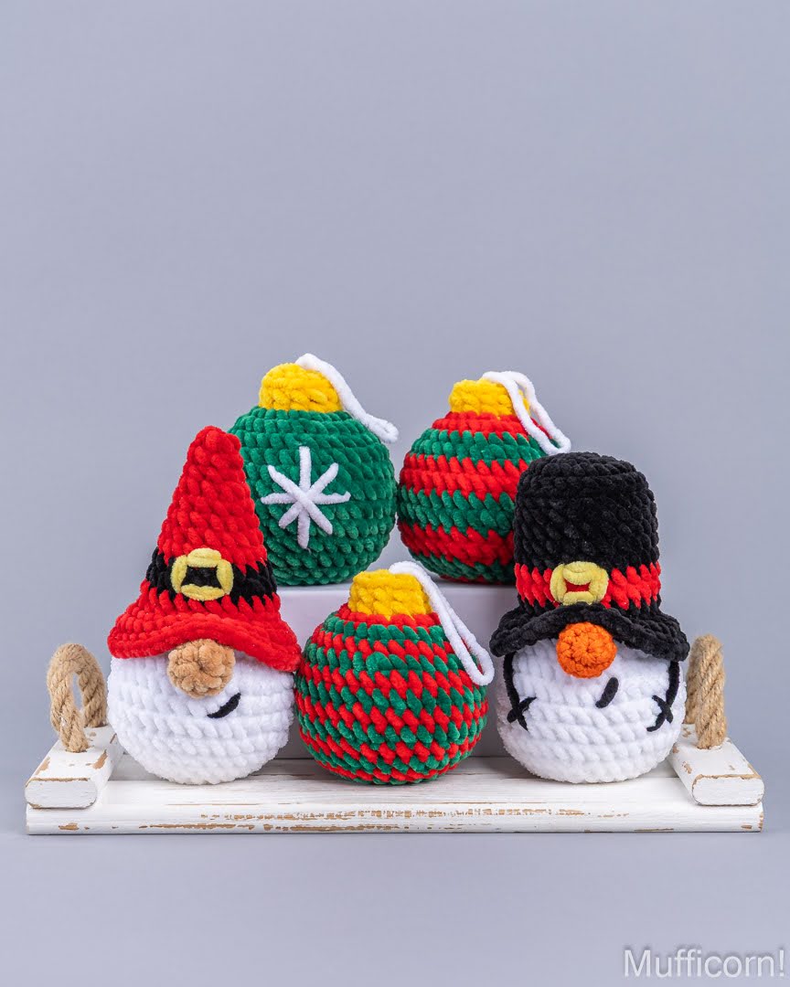 Five handmade crocheted Christmas ornaments displayed on a white wooden crate against a solid gray background. The ornaments include three bauble-shaped pieces and two gnome-shaped ornaments with red, green, white, black, and yellow yarn details.
