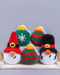 Four crocheted Christmas ornaments displayed on a white wooden surface against a gray background. The ornaments include a santa gnome with a red hat, a snowman with a black hat, and three striped green and red ornaments with yellow tops.