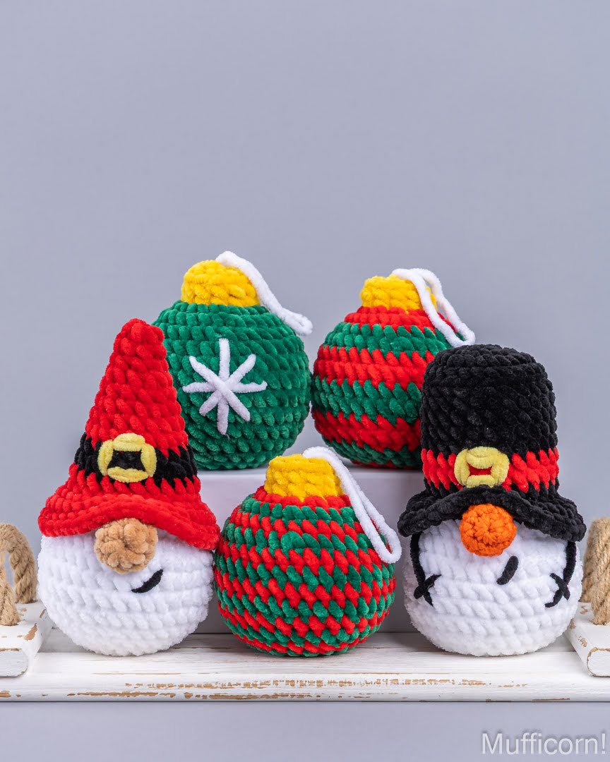 Four crocheted Christmas ornaments displayed on a white wooden surface against a gray background. The ornaments include a santa gnome with a red hat, a snowman with a black hat, and three striped green and red ornaments with yellow tops.