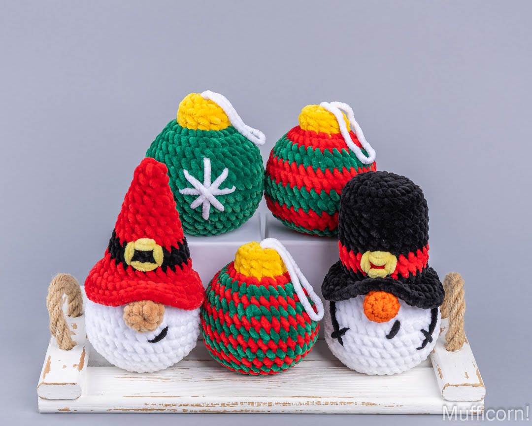 Five handmade crocheted Christmas ornaments displayed on a white wooden tray against a gray studio background. The ornaments include two gnome faces, two Christmas balls, and one ornament with a striped pattern, all in red, green, white, black, and yellow colors.