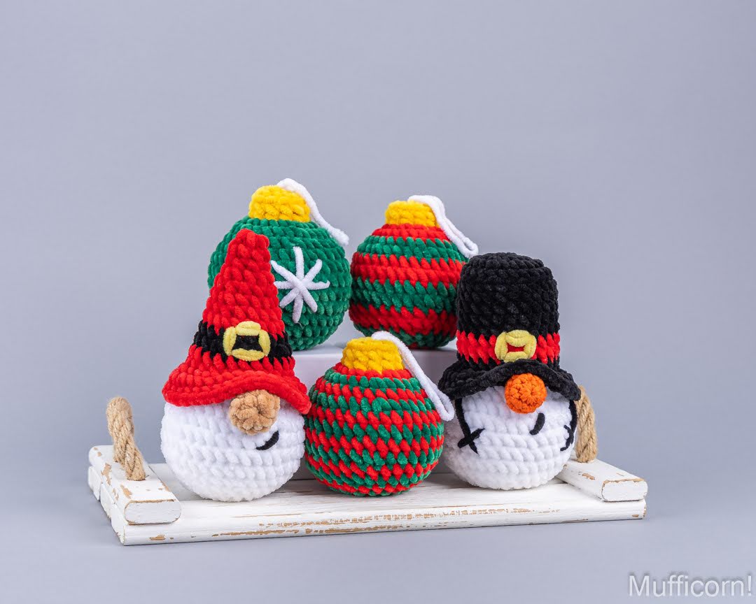 Four crocheted Christmas ornaments displayed on a small white wooden tray against a solid gray studio background. The ornaments include festive green and red baubles with yellow tops, a snowman head ornament in white, red, black, and orange, and a snowman ornament wearing a red hat with a white star on its green body.