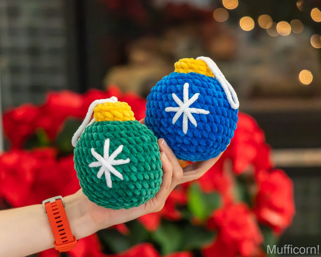 A person's hand holds two crocheted Christmas ornament balls, one green and one blue, each with a white star design, yellow top, and white hanging loop. The background is a blurred festive setting with red flowers and warm bokeh lights.